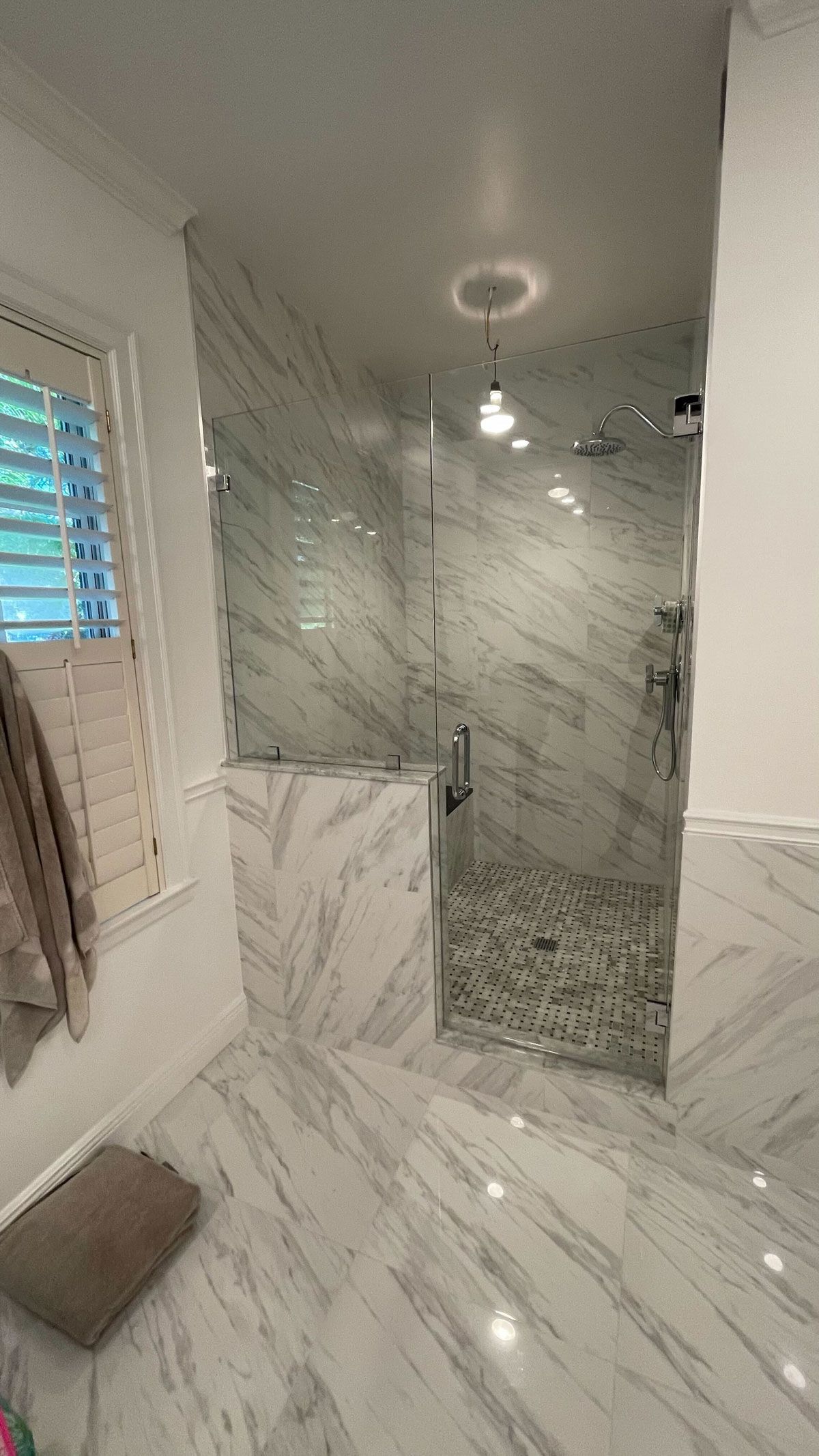 Frameless glass shower enclosure with a hinged door and chrome hardware in a marble-tiled bathroom, with a basketweave mosaic shower floor