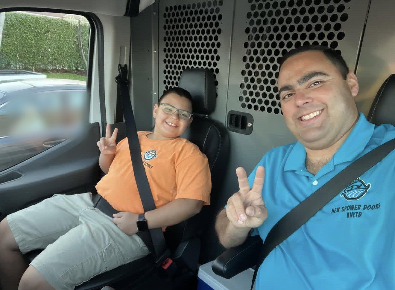 Alberto Alvarez and his son smiling and making peace signs in the front of the company van, Alberto in a turquoise New Shower Doors Unlimited polo and his son in an orange company T-shirt