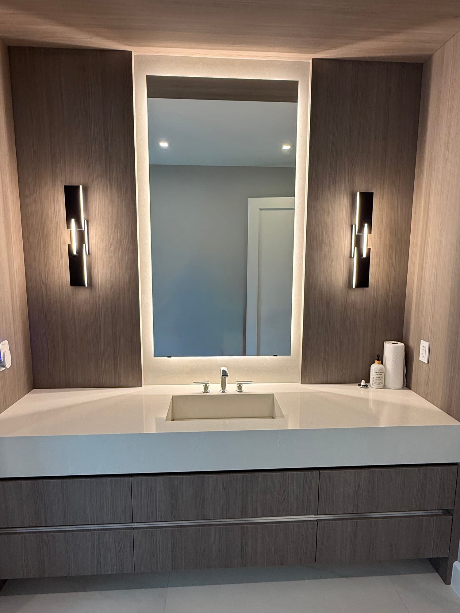 Backlit mirror set into a stone-clad recess between wood-panelled walls, its light spilling onto the wall above a floating vanity with an undermount basin