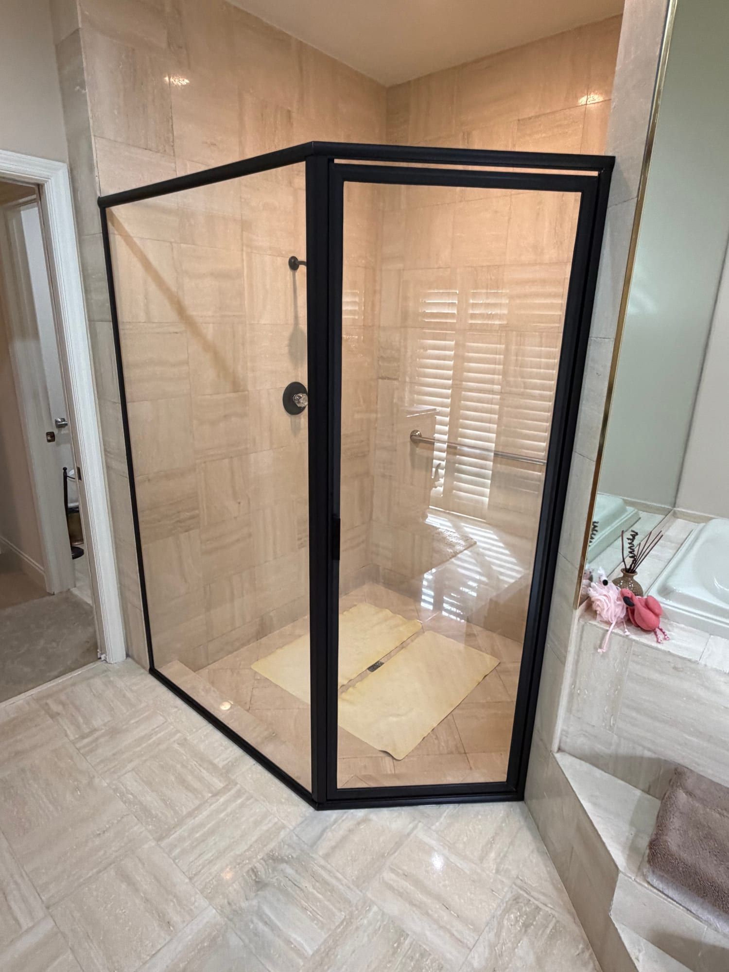 Neo-angle corner shower enclosure framed in matte black, its door and side panels set against travertine-look tile, the shower floor still papered from the install