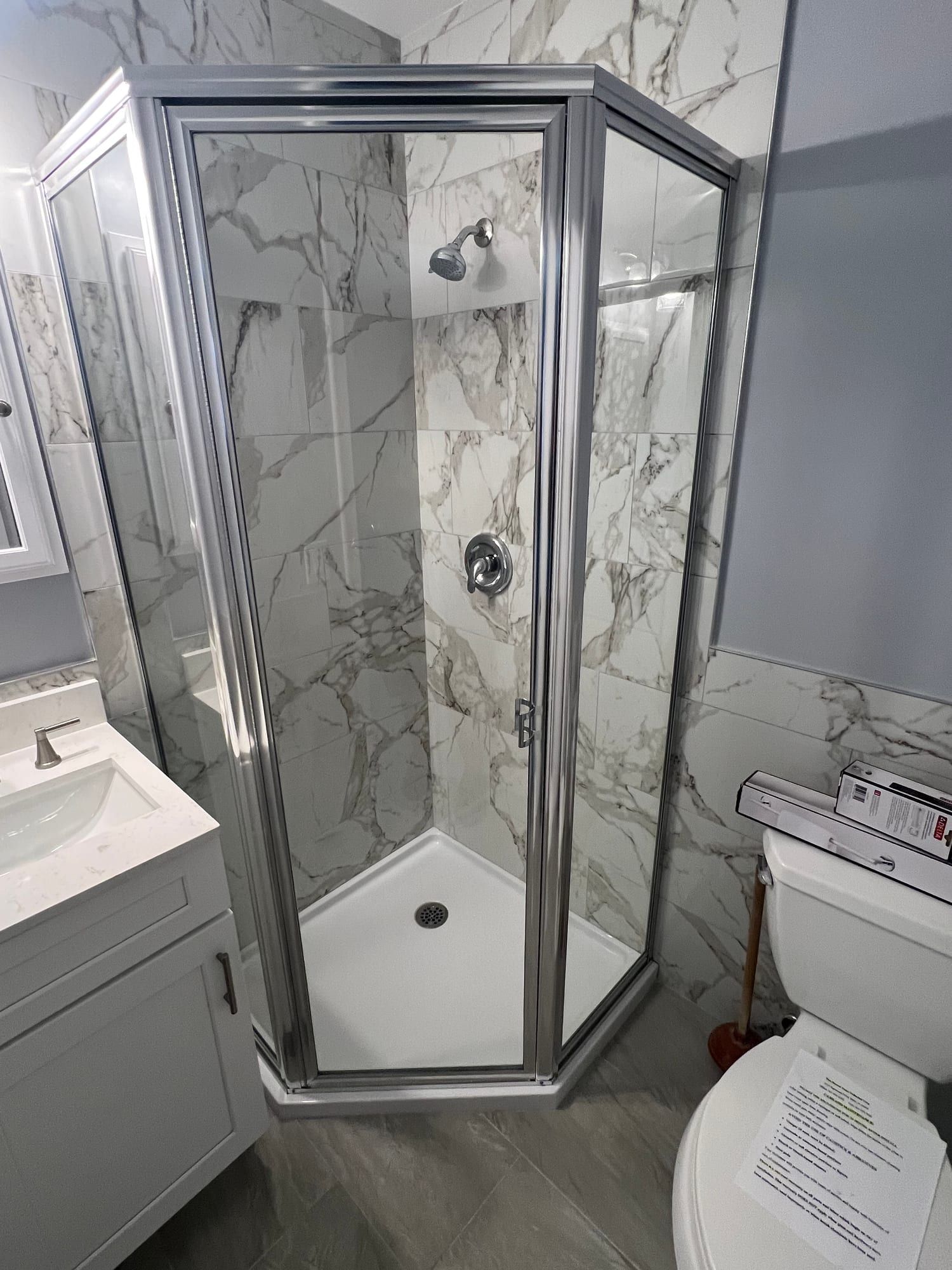 Neo-angle corner shower enclosure framed in polished chrome, with a hinged glass door, marble-look porcelain walls and a white shower base