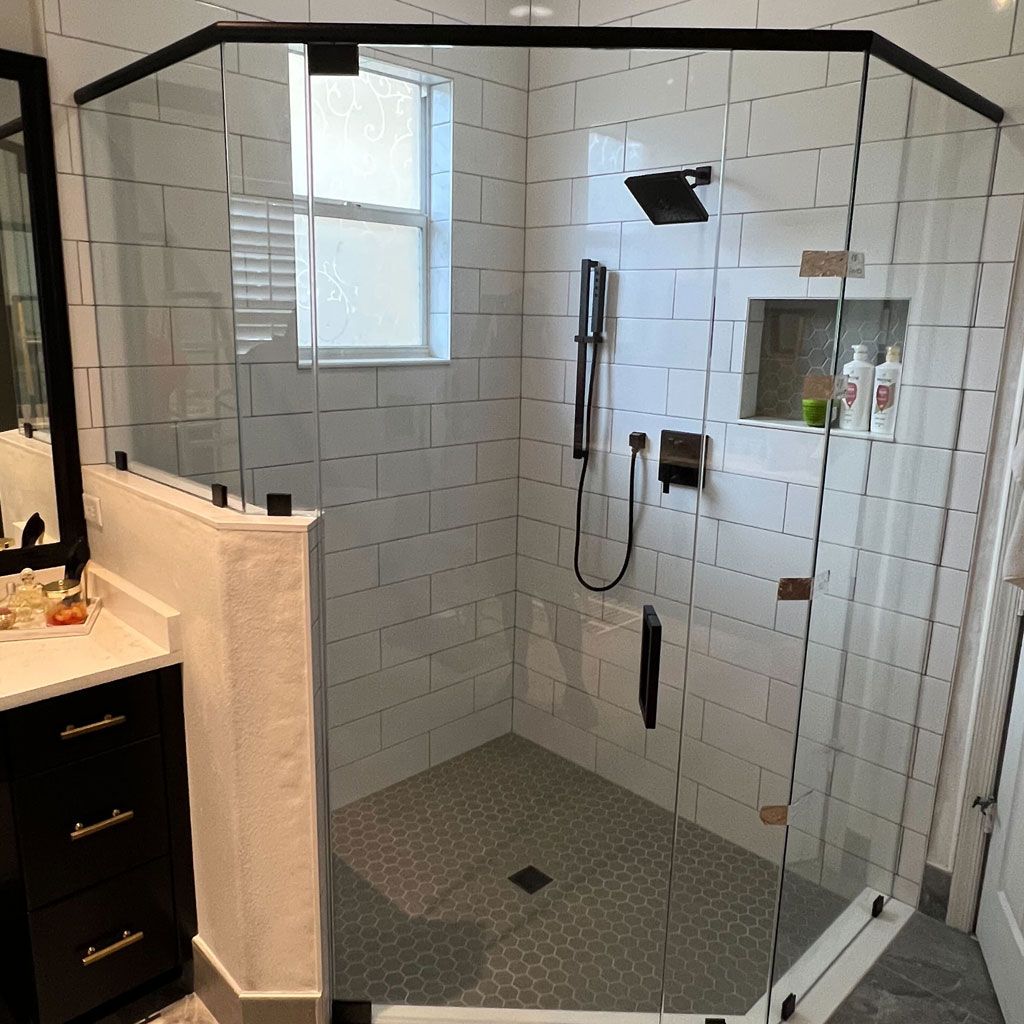 Neo-angle frameless glass shower enclosure with matte black clamps and support bar, white subway tile and a grey hexagon mosaic floor