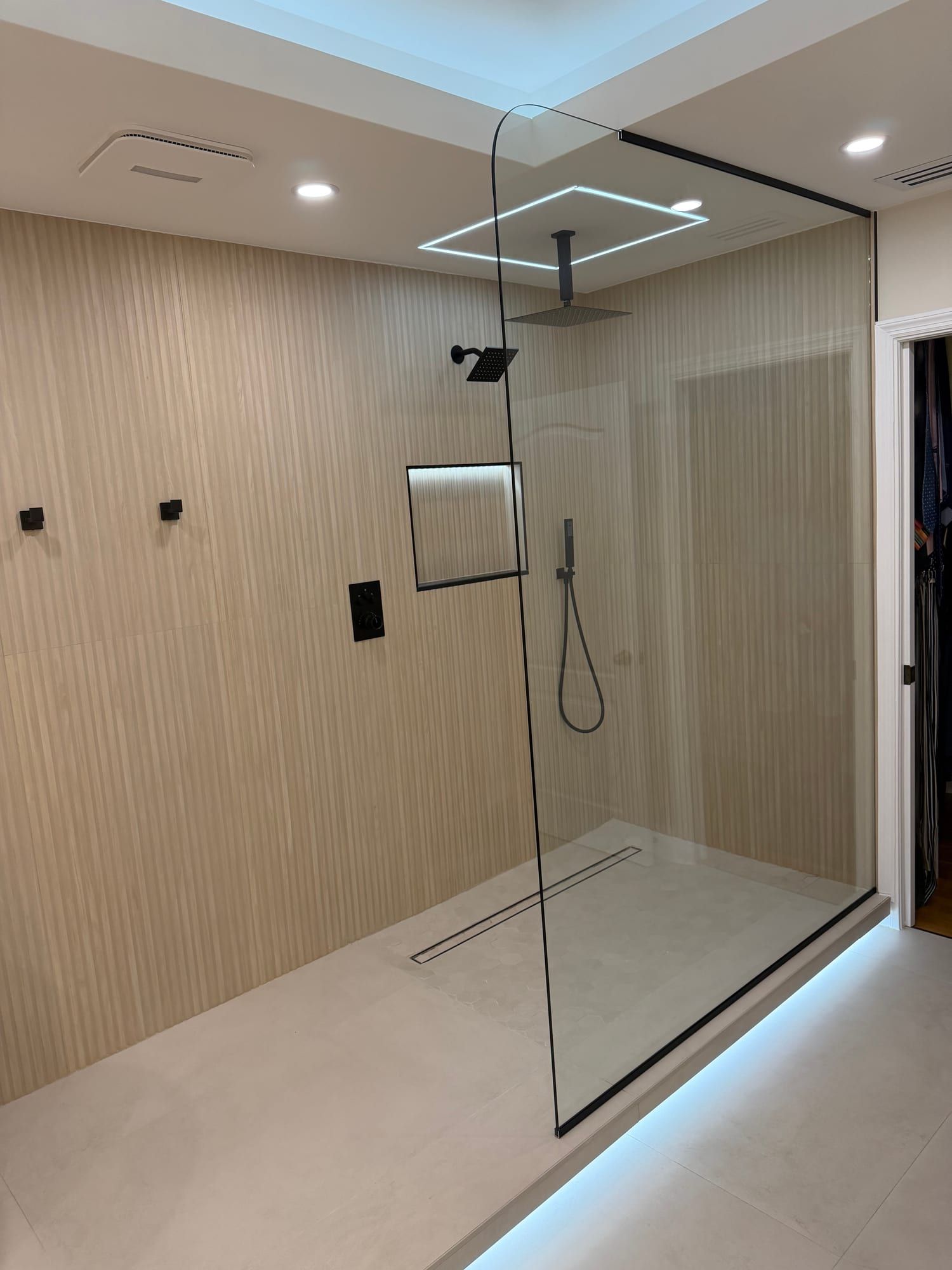 Frameless walk-in shower built without a door, its single fixed glass panel curved along the top edge and carried to a slim black channel at the ceiling, with a matte black ceiling rain head, wall head, hand shower and thermostatic valve, fluted beige tile, a lit recessed niche, a linear drain and LED light washing out from under the raised shower floor