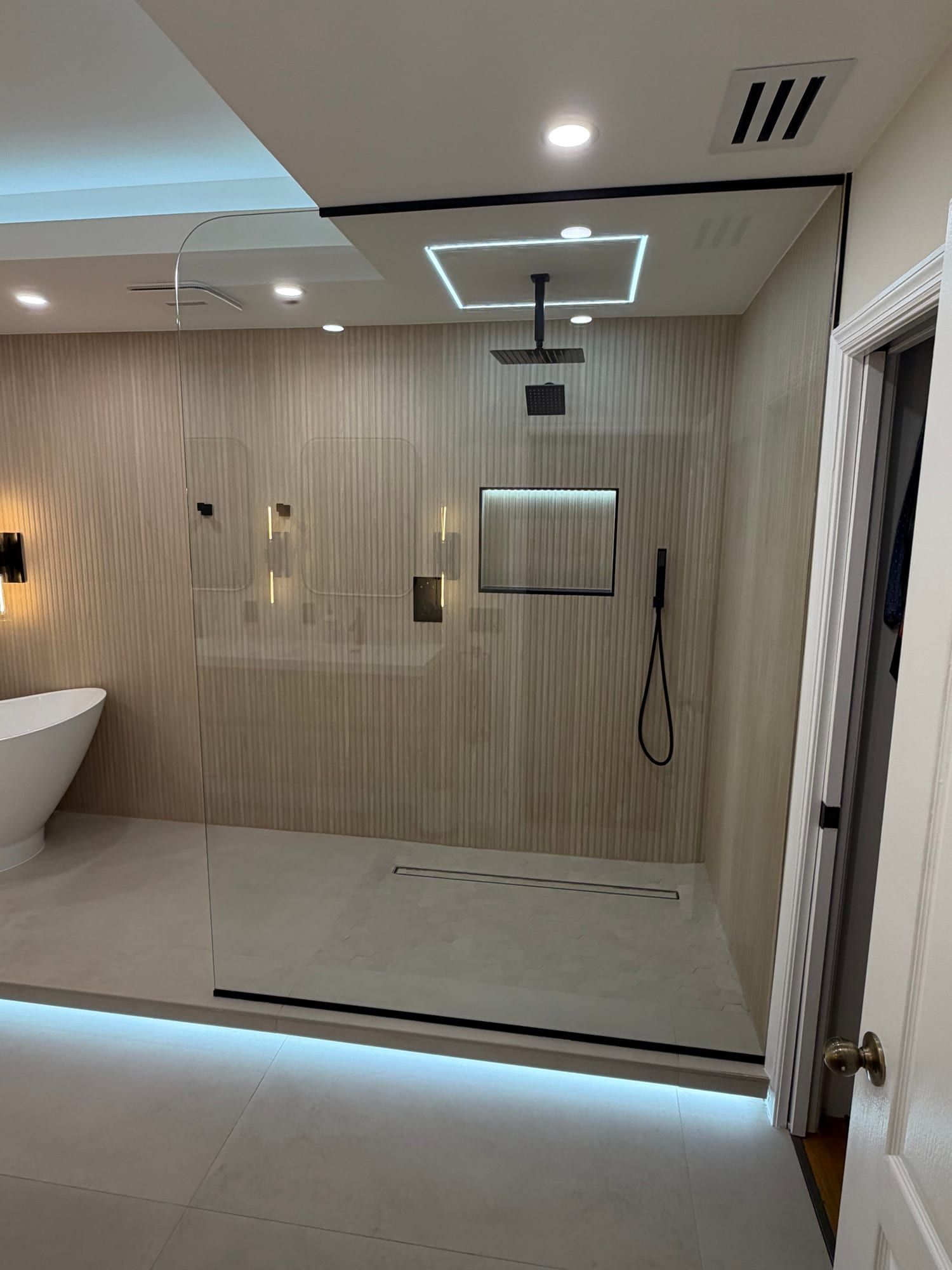 The same walk-in enclosure seen across the bathroom, its fixed glass panel spanning the full opening beside a freestanding tub, with a matte black ceiling rain head and hand shower, fluted beige tile, a lit recessed niche, a white hexagon mosaic shower floor and a linear drain