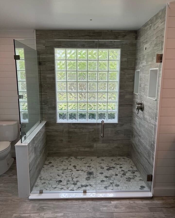 Frameless glass shower enclosure built around a glass block window, with wood-look tile walls and a marble hexagon mosaic floor