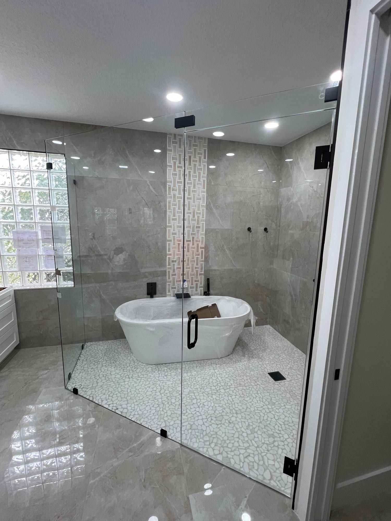 Frameless glass enclosure running floor to ceiling around a freestanding tub and shower, with matte black clamps, a black ladder pull, grey marble-look porcelain walls and a white pebble mosaic floor