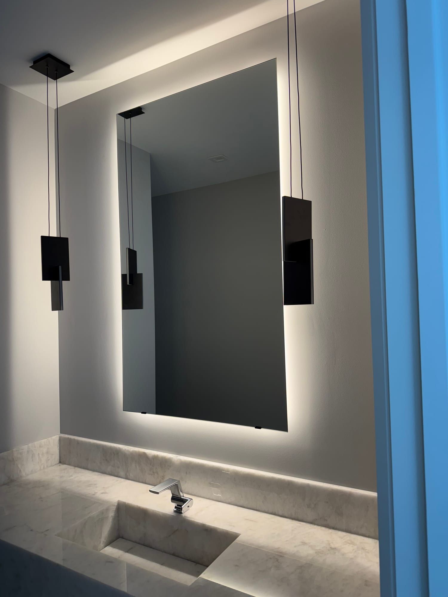 Frameless backlit mirror floating on the wall between two black pendant lights, its LED halo lighting the wall behind it above a marble vanity