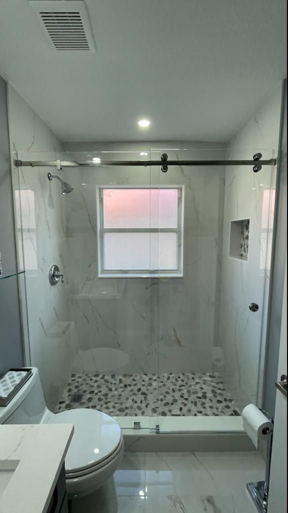 Sliding glass shower door on a chrome top track over a marble-tiled alcove, with a pebble mosaic shower floor