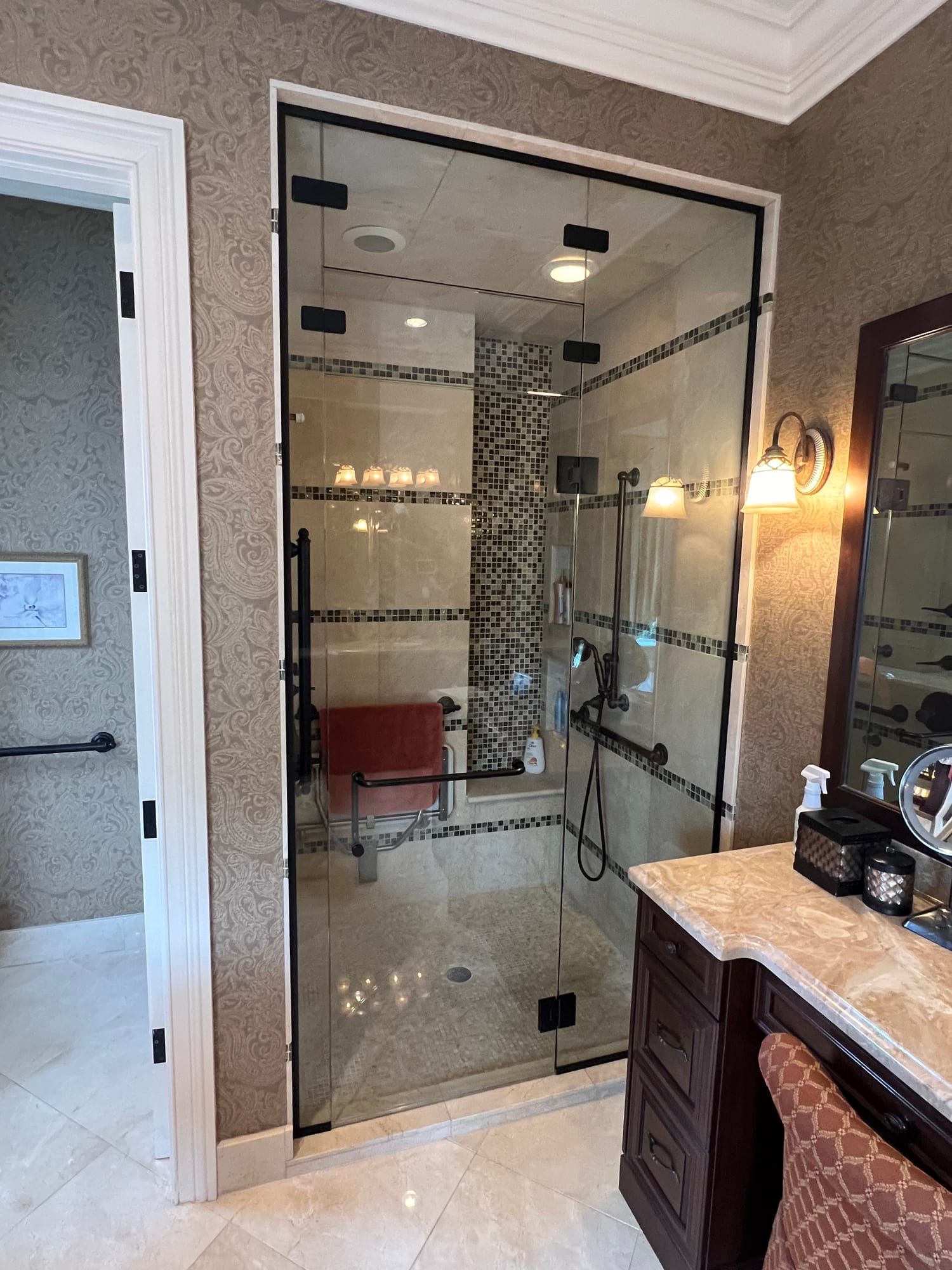 Steam shower enclosure sealed by a glass panel across the ceiling, its door and fixed panel held by dark bronze clamps, with travertine walls, a glass mosaic border and a built-in bench