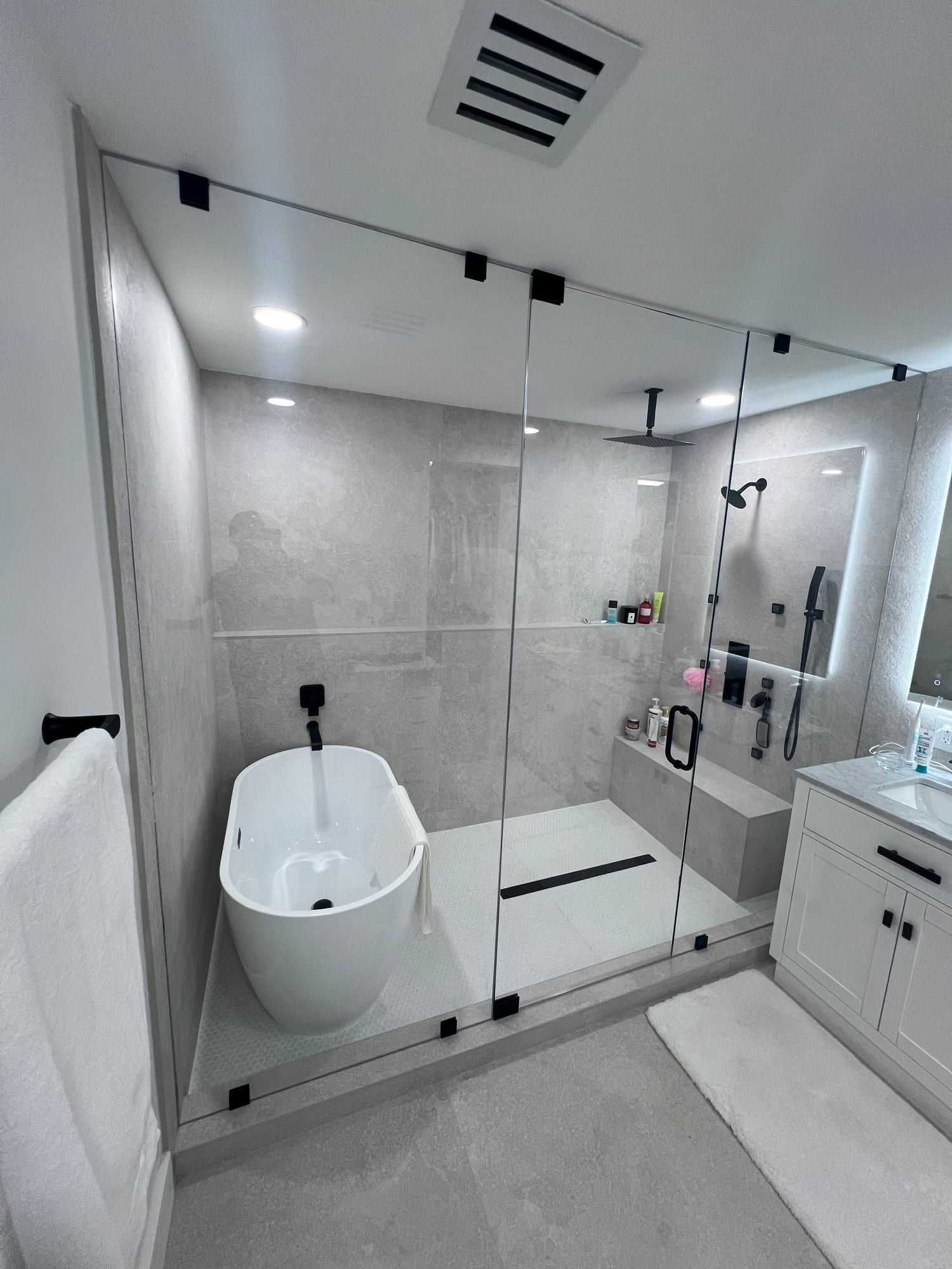 Steam shower enclosure with frameless glass carried to the ceiling on matte black clamps, holding a freestanding tub, a ceiling rain head, a built-in bench and a linear drain